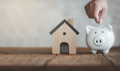 hand putting coin on White piggy bank with wooden house on wood table and blurred copy space background. Concept for financial home loan or money saving for house buying.