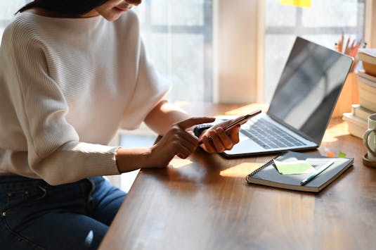 Cropped image of beautiful woman working as secretary holding/using a black blank screen smartphone while sitting in front her blank screen computer laptop over comfortable living room as background.